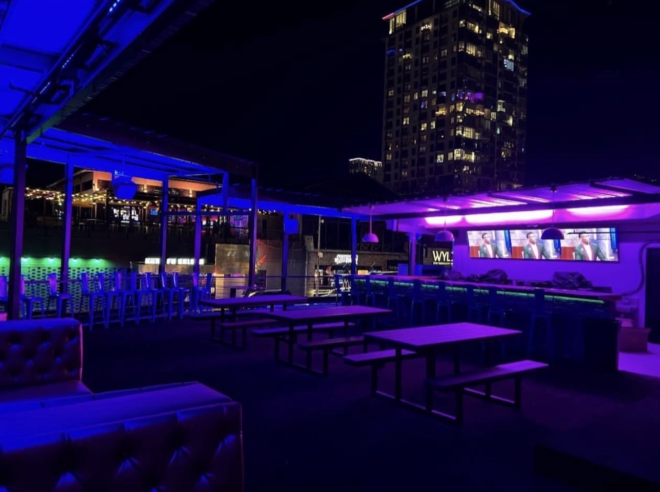 Willow's rooftop at night washed in blue light, with picnic tables, TV screens and a lit downtown tower behind
