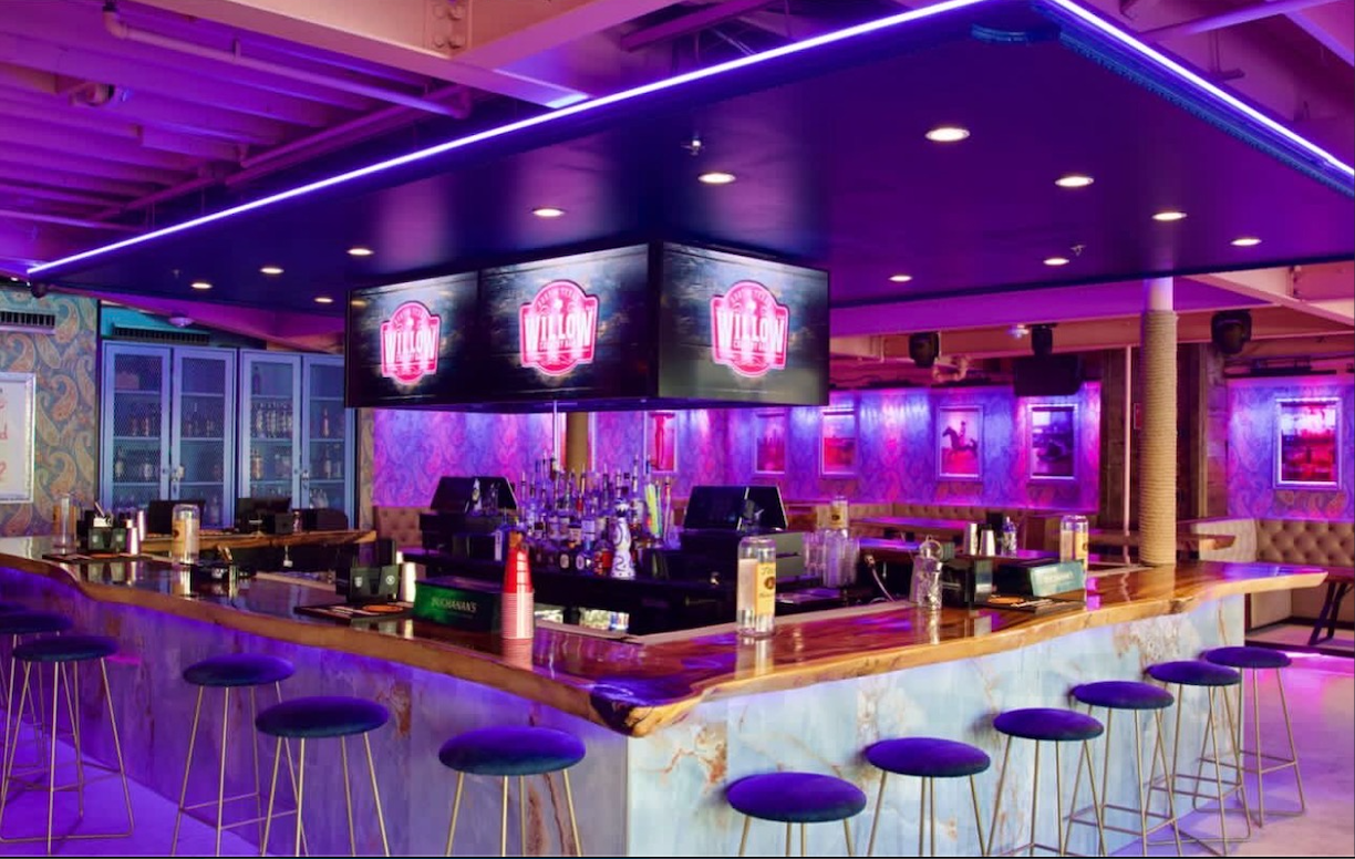 Willow's illuminated indoor bar with a glowing onyx base, blue velvet stools and WILLOW screens under purple light